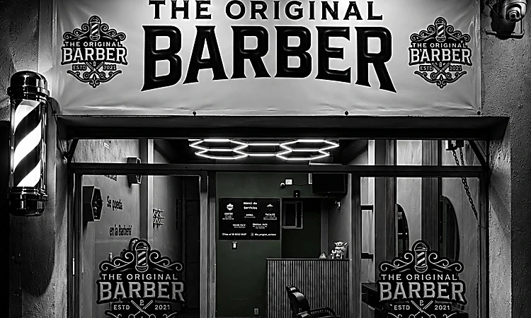 Fachada de The Original Barber: lona blanca con el nombre, poste de barbero a la izquierda y puerta de vidrio con las insignias