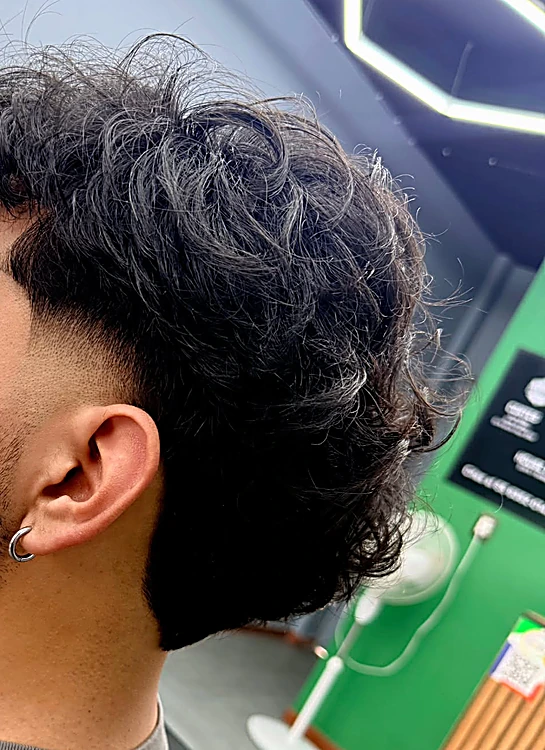 Fade bajo con rizos arriba, visto de perfil; atrás se alcanza a ver el panal de luz y el muro verde